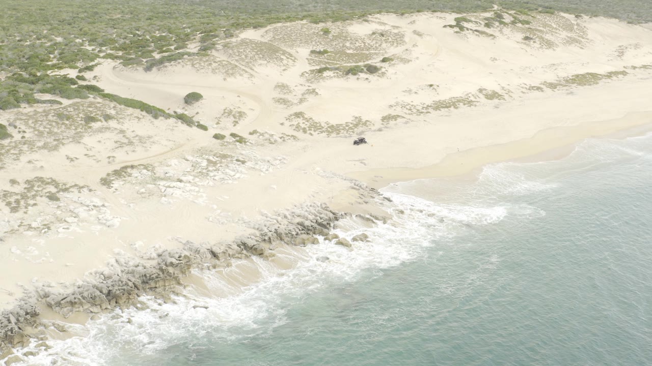 Drone view over breaking surf and rocky sand with a small off-road vehicle near the upper beach, surrounded by pale dunes and sparse green plants, with no visible people.