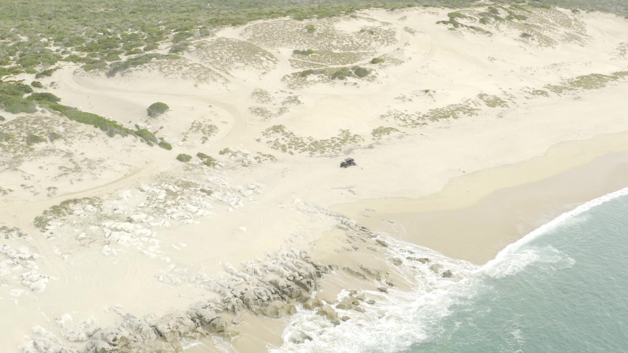 At 0:00, a high aerial view shows a small dark UTV on pale beach sand near low dunes, scattered scrub, rocks, and blue-green surf, with no visible people.