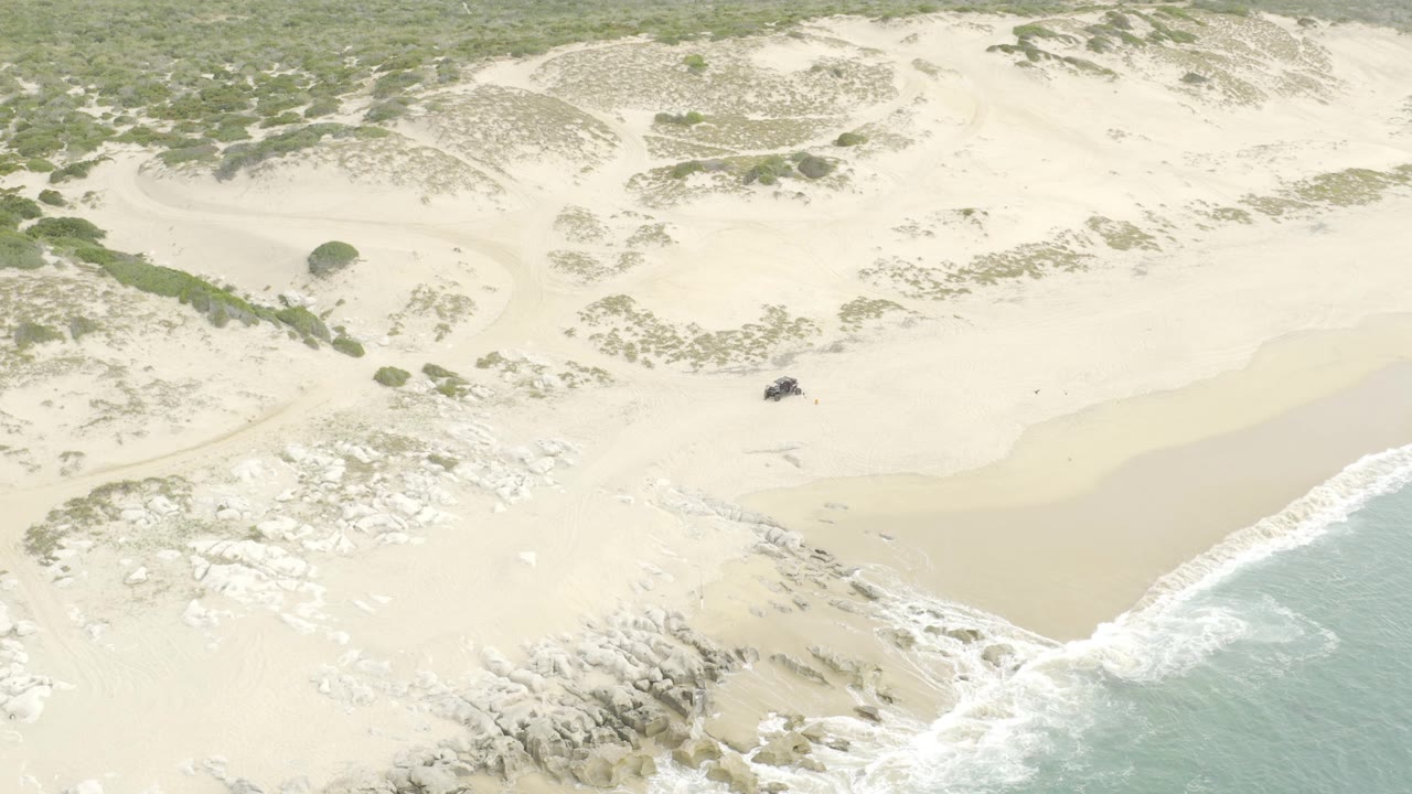 At 0:02, pale dunes and patchy green vegetation fill the frame while the dark UTV remains on the open beach near the surf line, with no visible people.