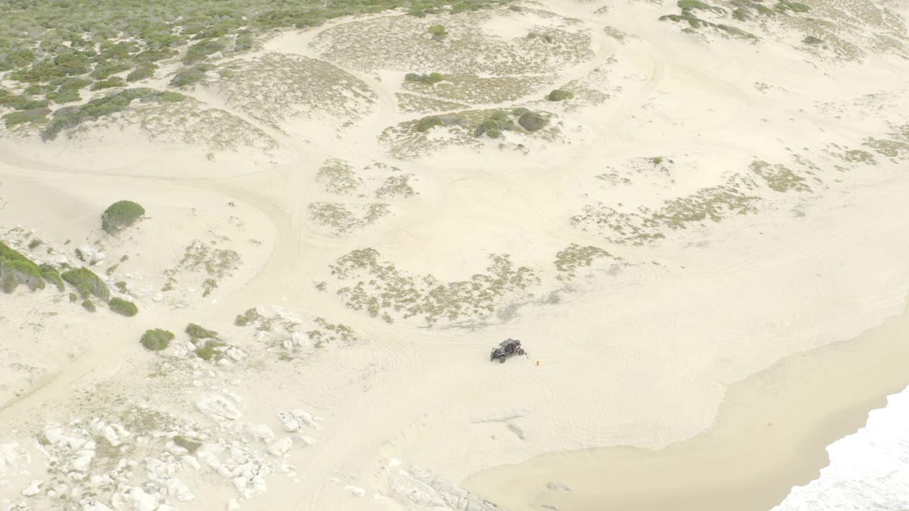 Overhead view of a dark UTV on pale sand dunes near vegetation and surf.