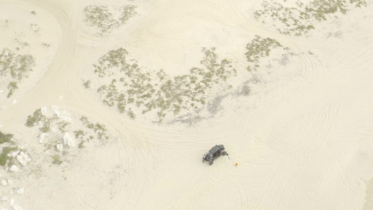 Top-down view of a dark off-road vehicle on pale sand near scrub vegetation.