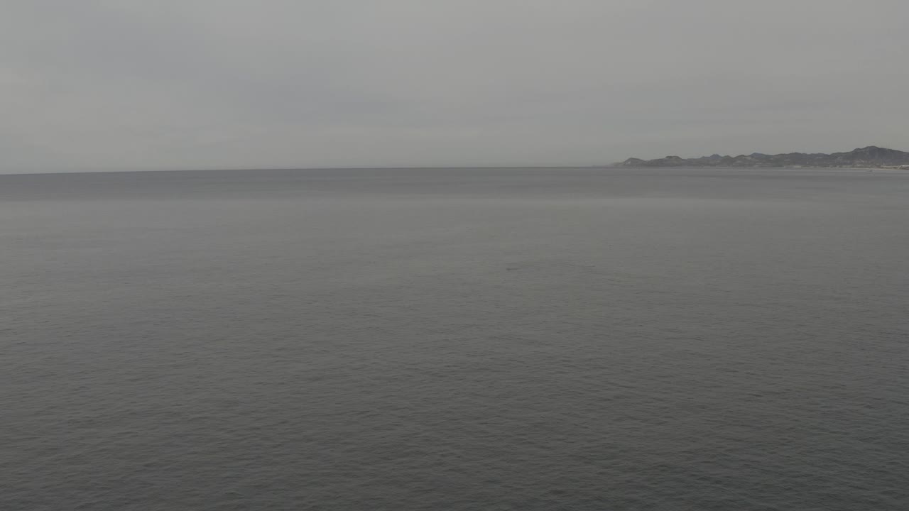 Mid-clip frame with calm grey sea in the foreground, distant coastline across the horizon, cloudy sky above, and no visible people.