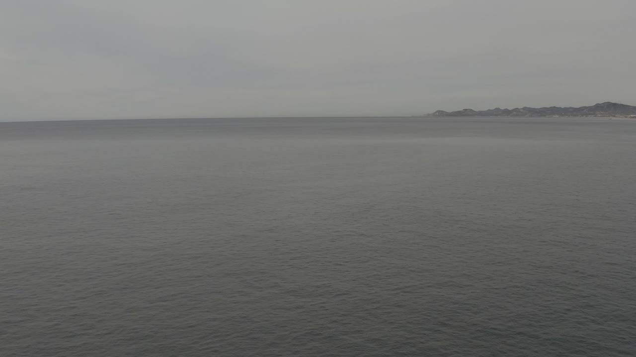 A wide aerial view shows dark grey ocean, a soft level horizon, faint land at the far right, and no visible people at the opening moment.
