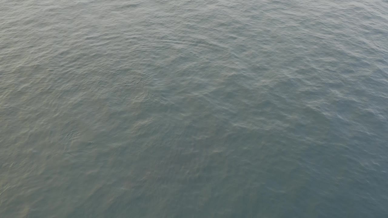 Mid-clip overhead view of calm ocean surface, with pale ripple streaks crossing darker blue-green water and no visible people.