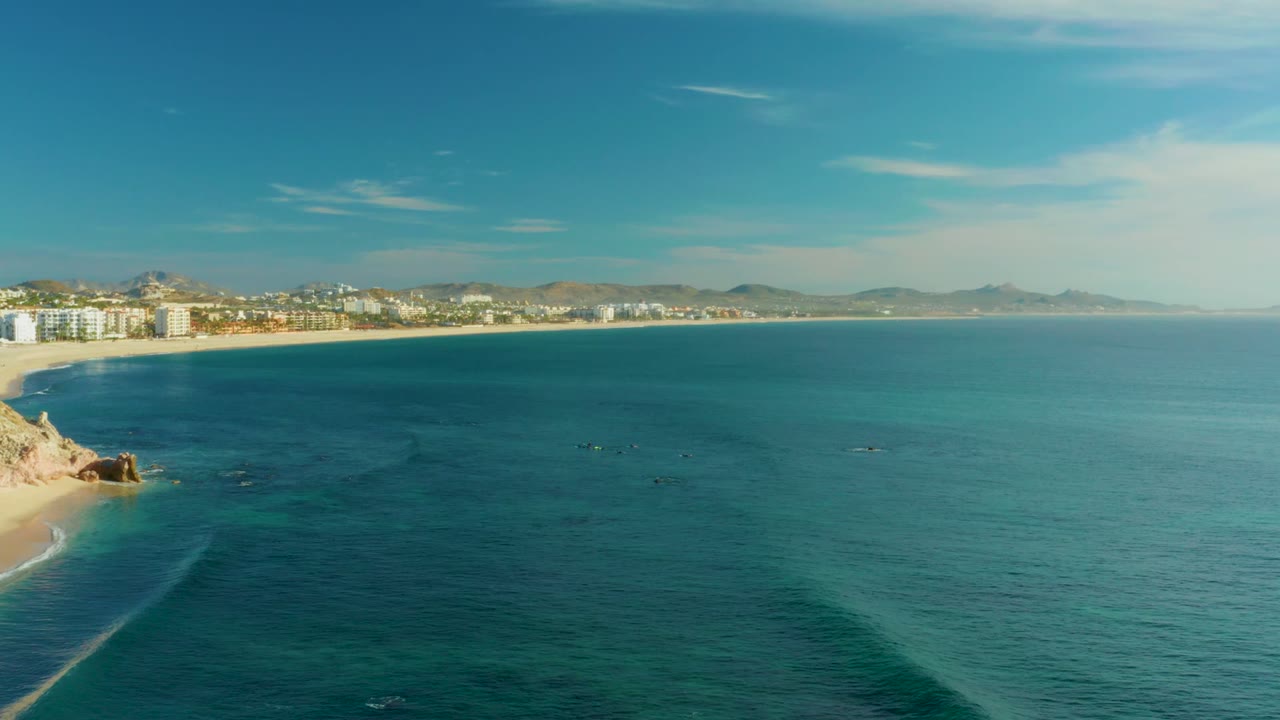 Wide aerial view at Playa Acapulquito, Los Cabos, with deep blue ocean filling the frame, a sandy beach and rocky point on the left, shoreline buildings in the distance, and no visible people.