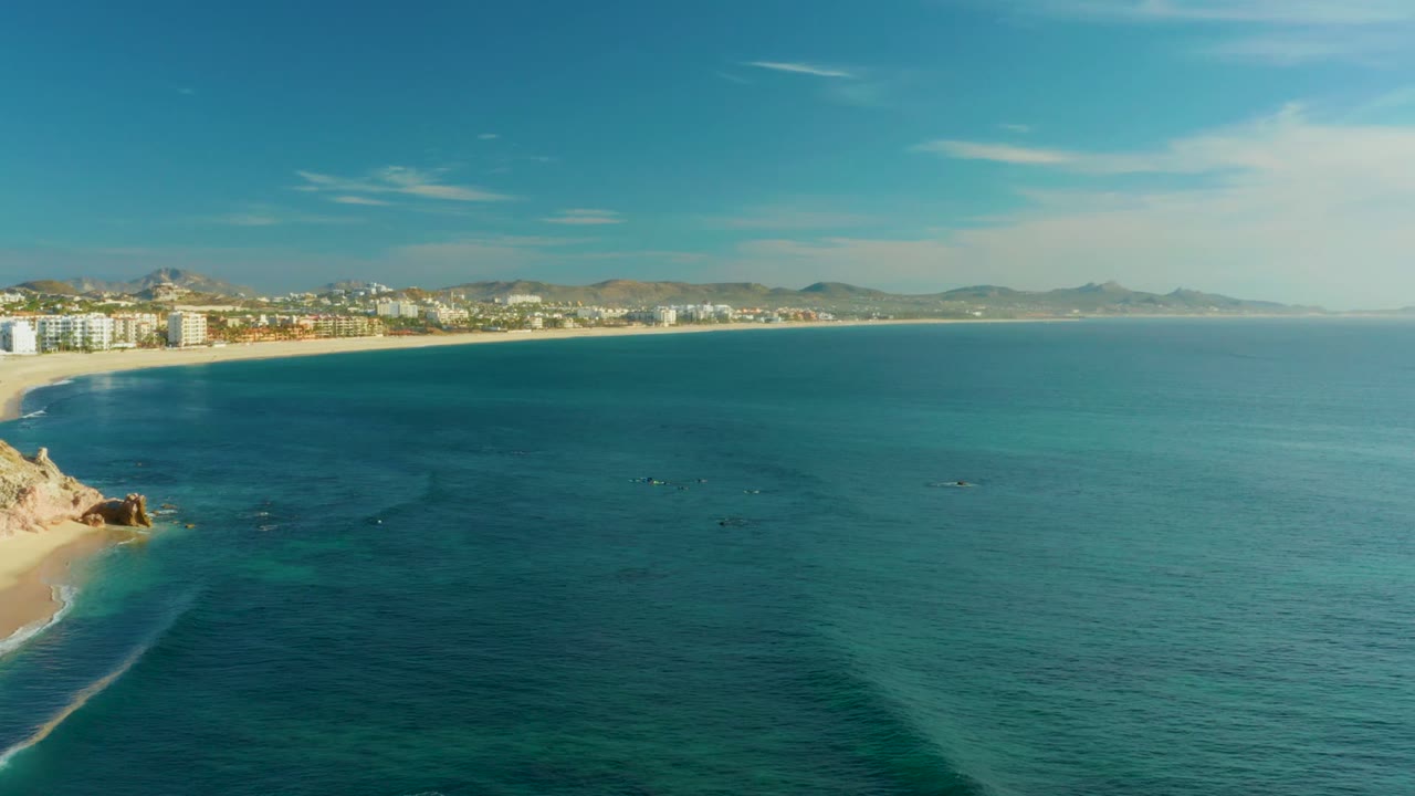 Aerial coastline view with clear water, beach, rocks, buildings, and hills.