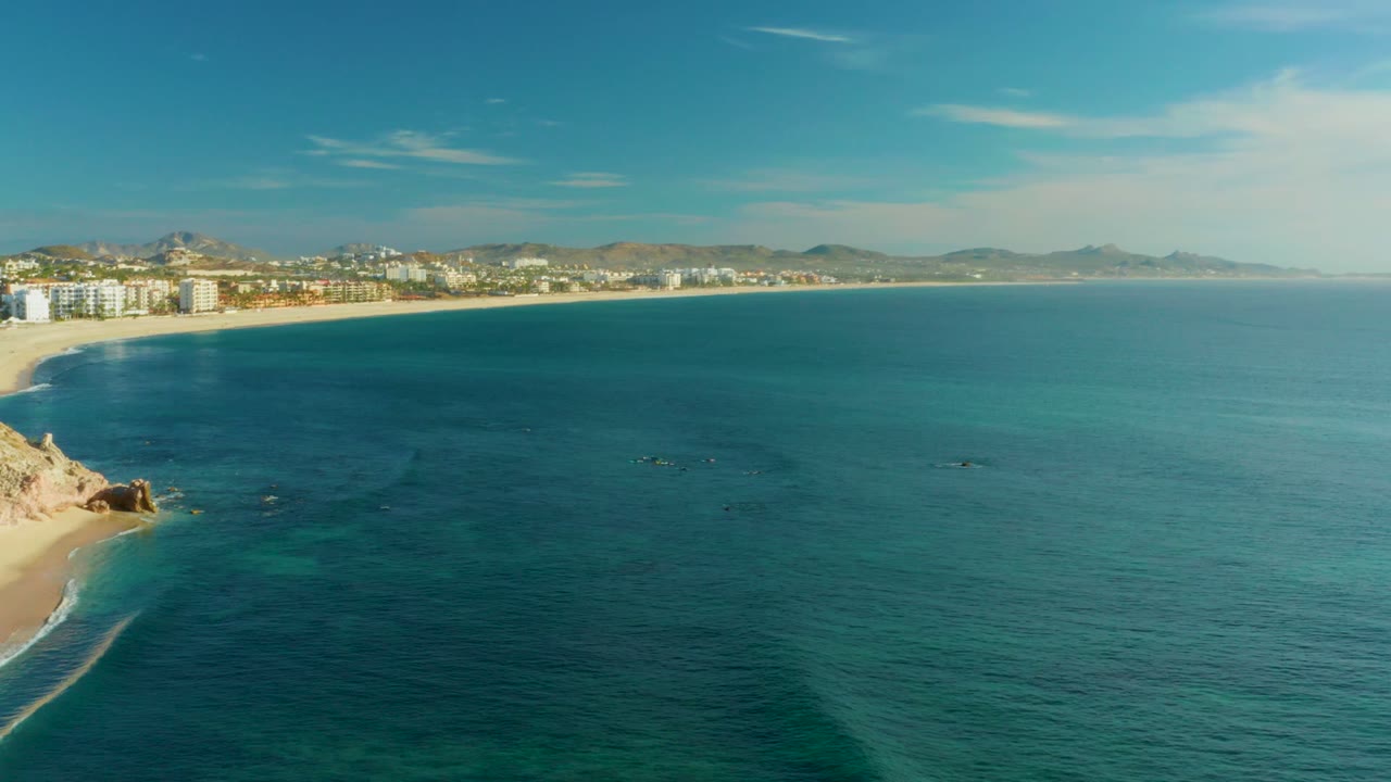 High aerial view at Playa Acapulquito with the rocky beach edge low left, a wide field of calm turquoise water, beachfront buildings along the upper shore, hazy hills beyond, and no visible people.