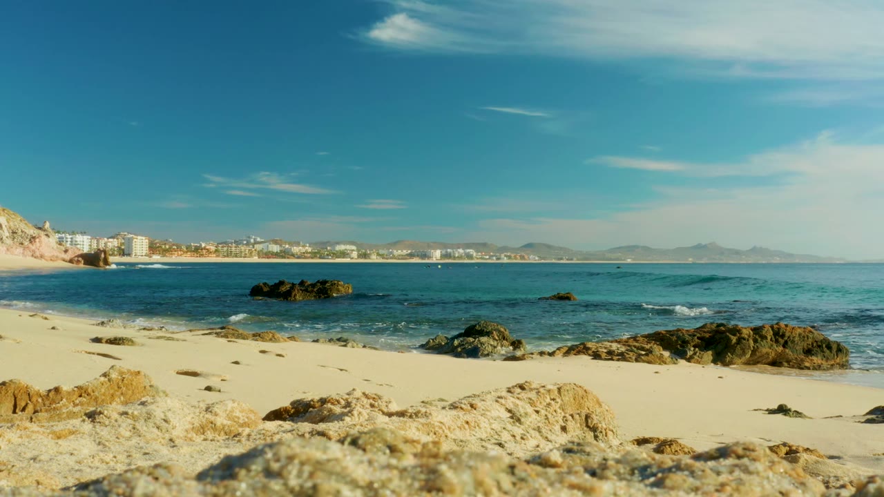 Rocky sandy shore with gentle waves and distant coastal buildings.