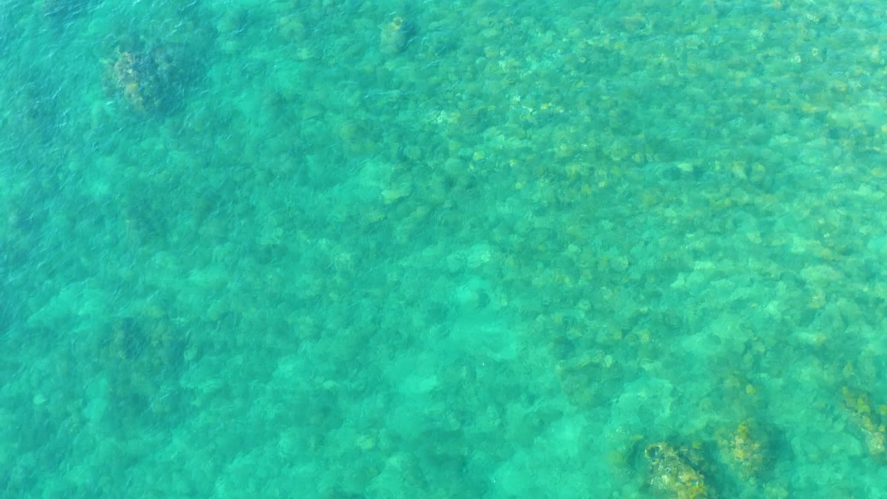 Clear turquoise water at Playa Acapulquito in Los Cabos reveals scattered underwater rocks near the upper and lower frame, with no visible people.