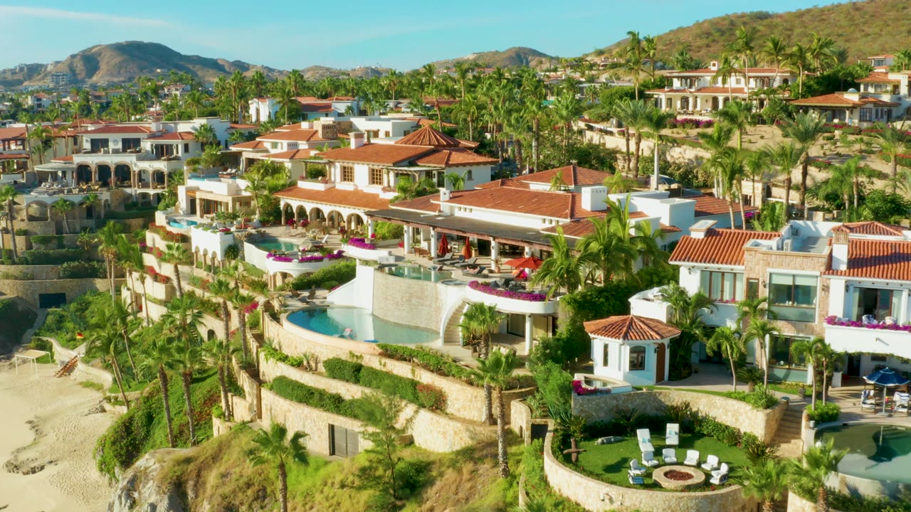 Aerial frame over Playa Acapulquito in Los Cabos showing red-roofed resort villas, palm trees, curved pools, sandy beach at left, hills in the background, and a small person on the sand.
