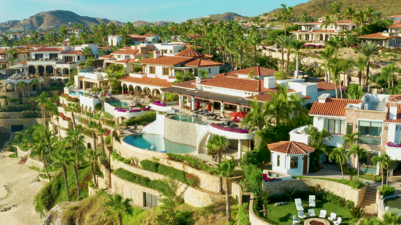 Aerial resort view with red roofs, pools, palm trees, sand, and distant hills.