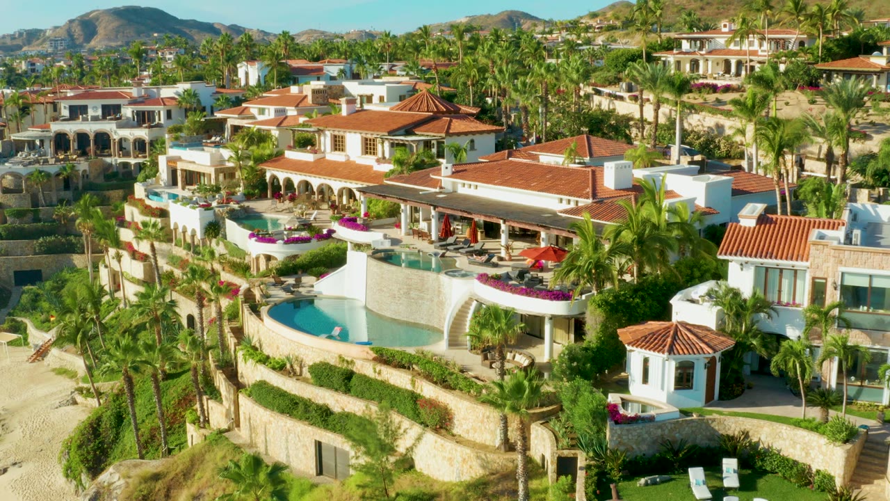 Playa Acapulquito resort seen from above with a curved pool in the lower left, white buildings and red roofs across the center, dense palms, bright terraces, and no visible people.