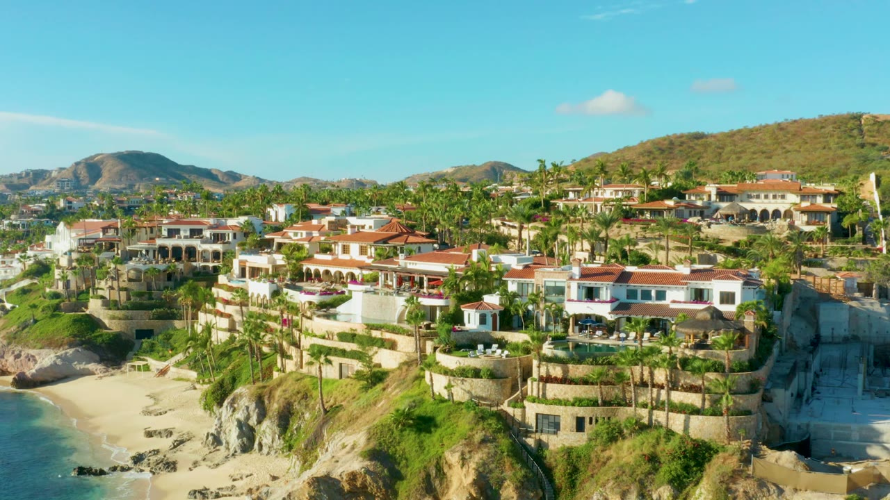 Opening aerial frame of Playa Acapulquito, Los Cabos, showing red-roofed coastal homes above a sandy beach, blue ocean at left, palms, dry hills, and no visible people.