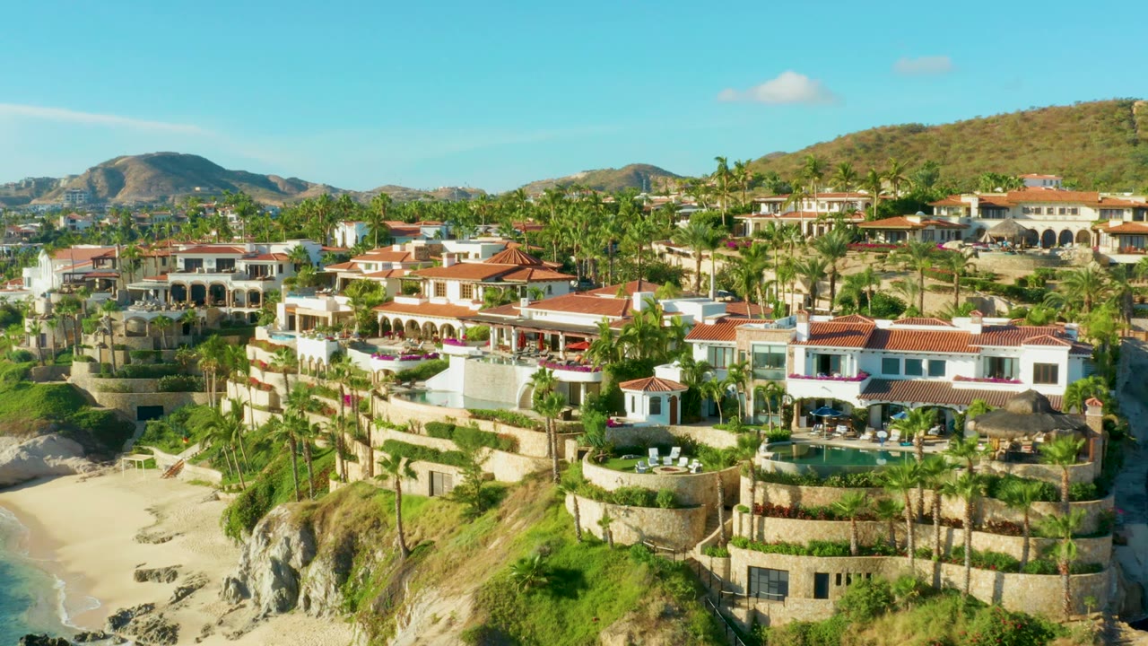 High oblique frame of Playa Acapulquito homes with red roofs and white walls, a sandy cove below, palms around the buildings, brown hills behind, and no visible people.