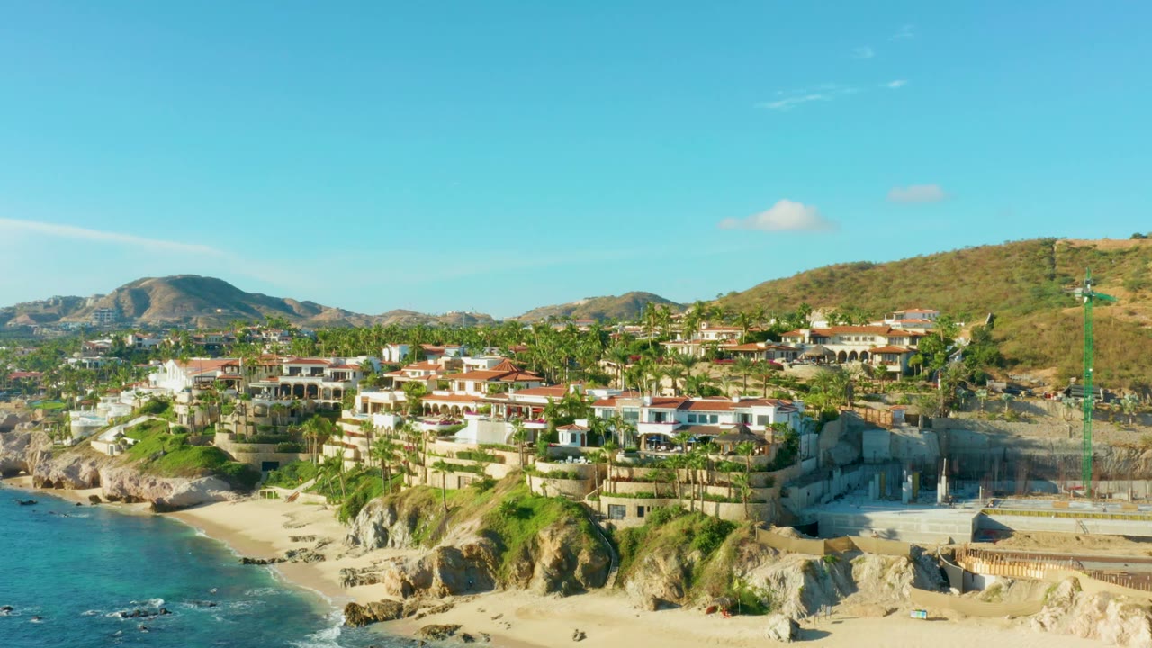 Aerial view of Playa Acapulquito, Los Cabos with turquoise water at left, a sandy beach below, hillside homes and palms across the center, a construction site at right, and no visible people.