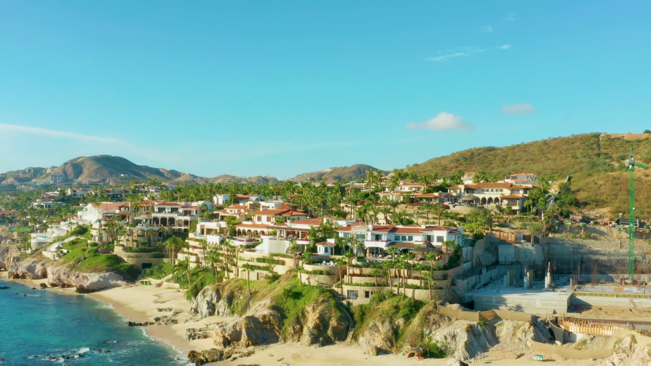 Coastal aerial with ocean, sand, palm-lined homes, hills, and a crane near construction.