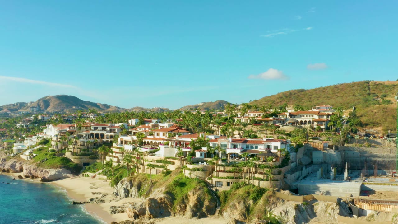 Aerial frame over Playa Acapulquito with more of the hillside neighborhood visible, turquoise water along the lower left, red-roofed buildings and palms centered, construction at right, and no visible people.