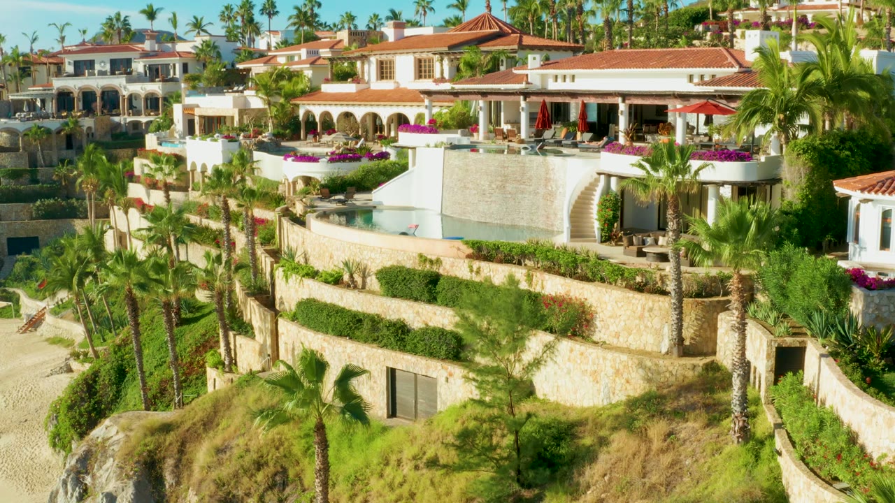 Elevated view of a Playa Acapulquito resort hillside with white buildings, red tile roofs, palm trees, stone terraces, flowering planters, and no visible people.