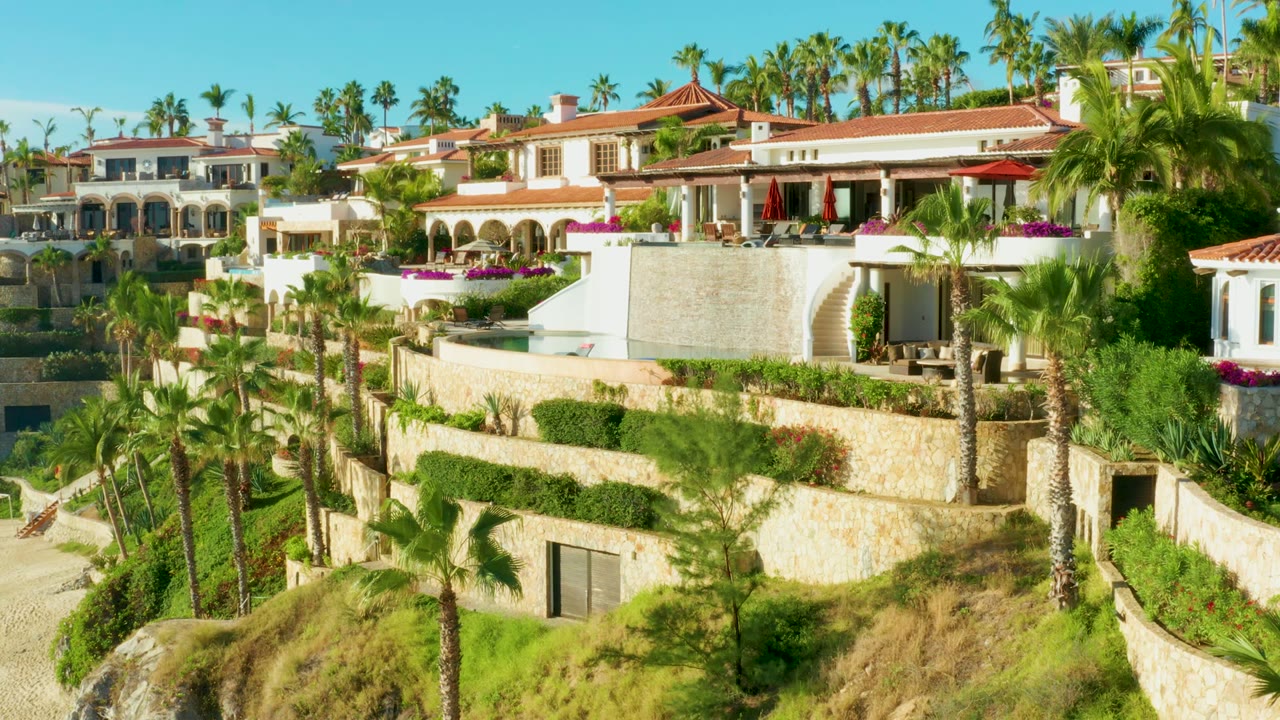 Elevated Playa Acapulquito resort frame with tall palms in front of red-roof villas, layered stone garden walls, bright green shrubs, and no visible people.