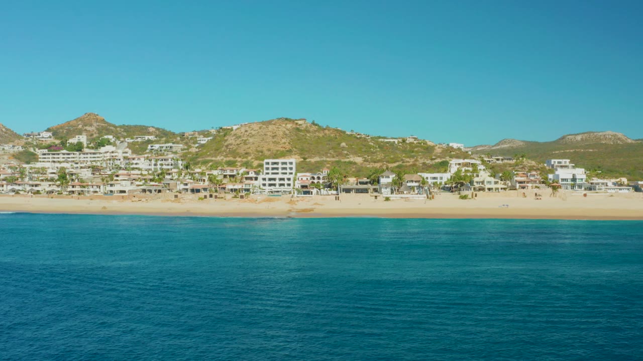 Wide view of Buzzards Beach in East Cape, Baja California Sur, with turquoise ocean foreground, pale sand, white hillside buildings, dry hills, clear blue sky, and no visible people.
