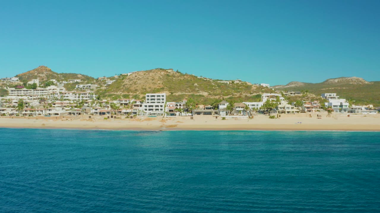 East Cape beach view showing a wide turquoise water foreground, sandy shoreline, white multi-story buildings and homes at the base of dry hills, with clear sky and no visible people.