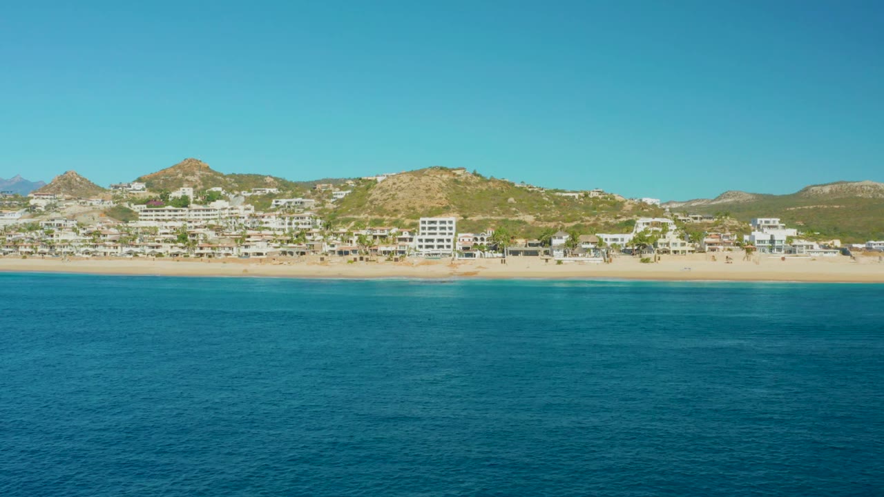 Opening frame of Buzzards Beach in Baja California Sur, with turquoise water in front, a sandy shoreline, low buildings, dry hills, clear blue sky, and no visible people.