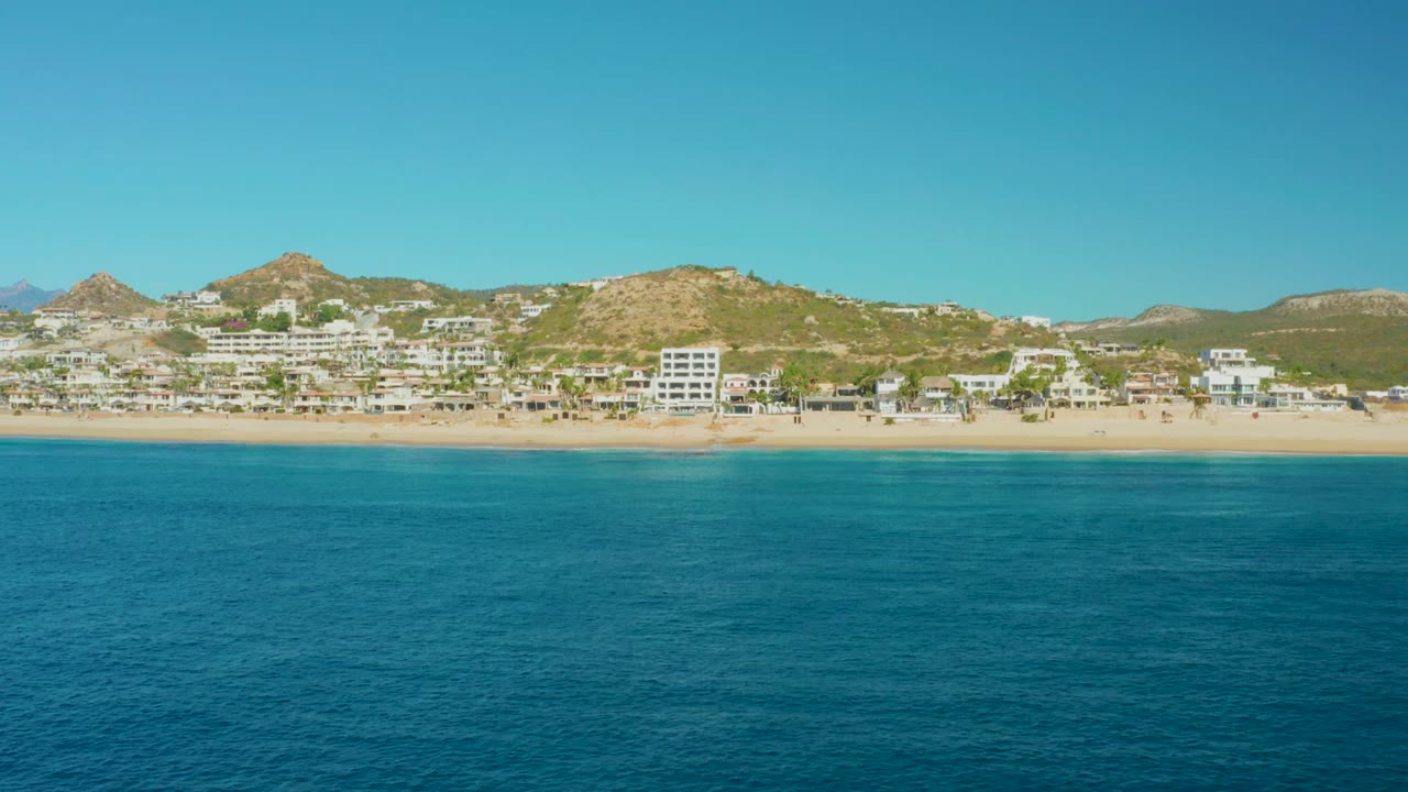 Wide coastal view with ocean texture, sandy shore, buildings, and low mountains.