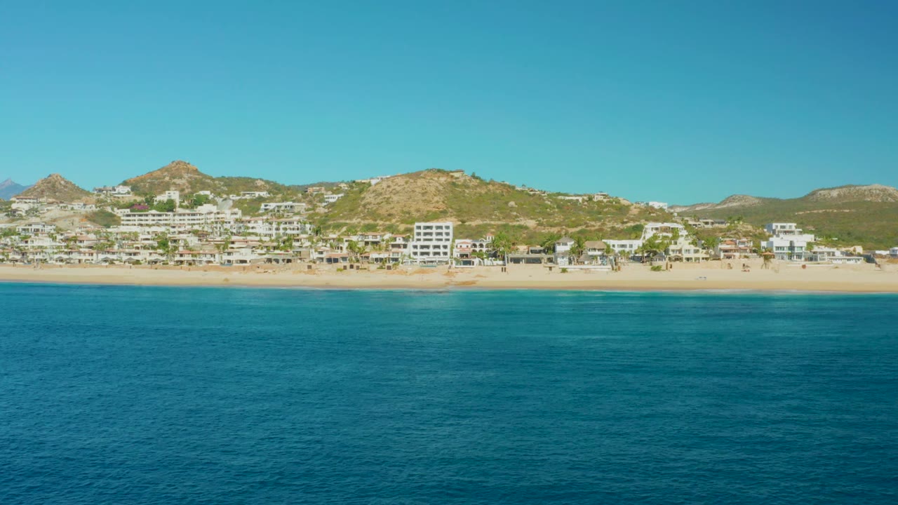 At 0:02, the Buzzards Beach shoreline remains broad across the frame, with blue-green water below, pale sand, small coastal structures, brown-green hills, and no visible people.