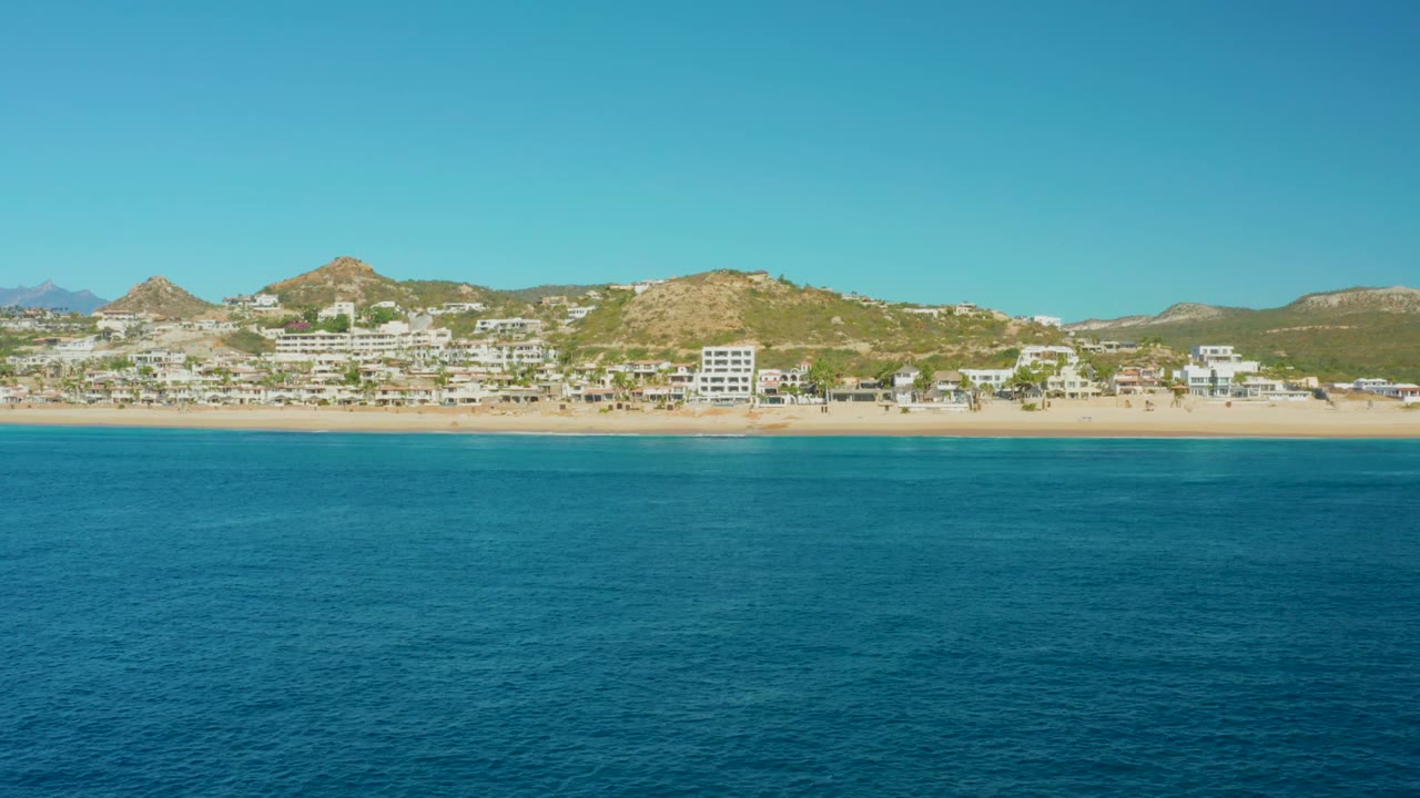 Opening view of Buzzards Beach in East Cape, Baja California Sur, with blue ocean in the foreground, a sandy shore, resort buildings, low hills, clear sky, and no visible people.