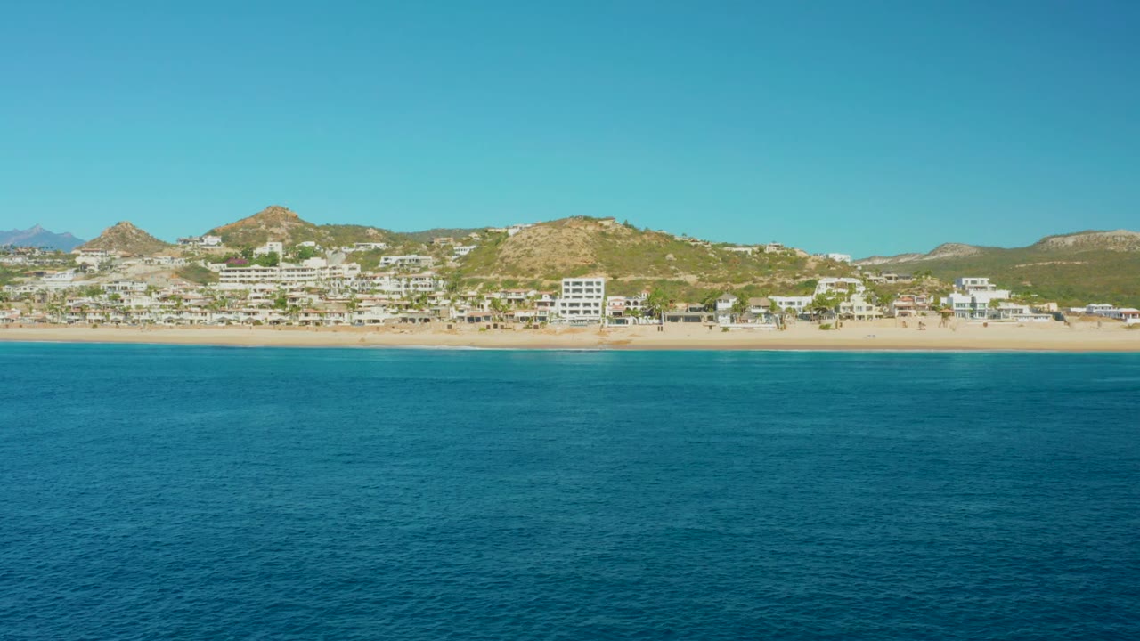 Calm ocean water in front of a beach lined with buildings and dry hills.