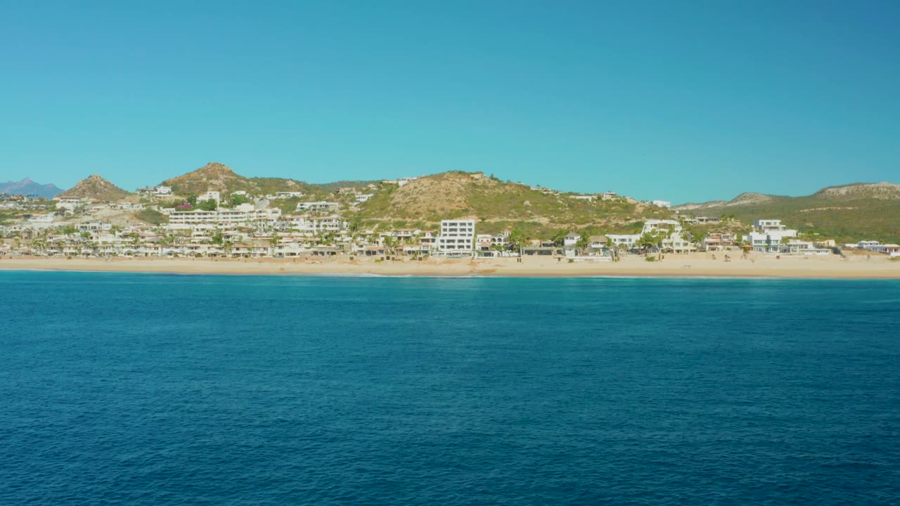 A wide view over the East Cape water shows gentle surface texture, a narrow sandy beach, clustered resort buildings, low hills, a cloudless sky, and no visible people.
