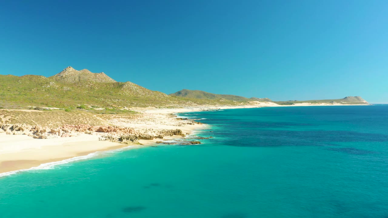 Opening frame of Buzzards Beach East Cape in Baja California Sur, with turquoise water, pale sand, rocky outcrops, green hills, blue sky, and no visible people.