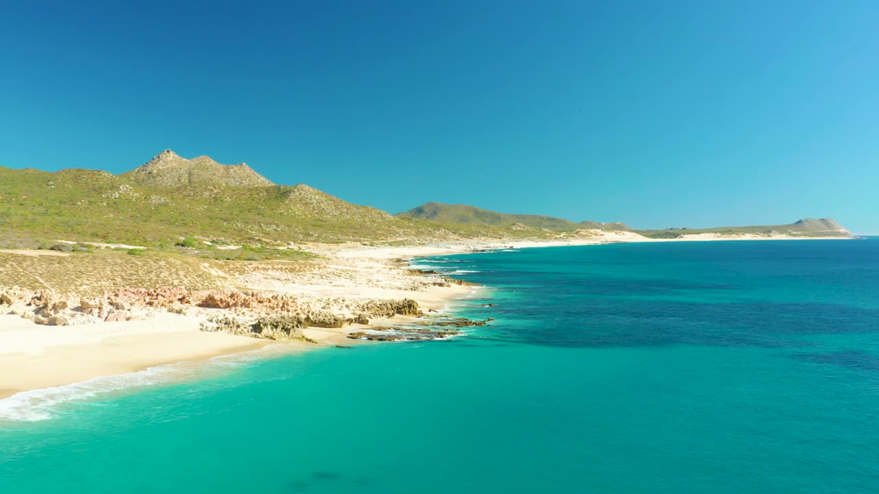 At 0:02, the Buzzards Beach shoreline shows pale sand and rocks on the left, deeper blue water to the right, low green hills on the horizon, and no visible people.