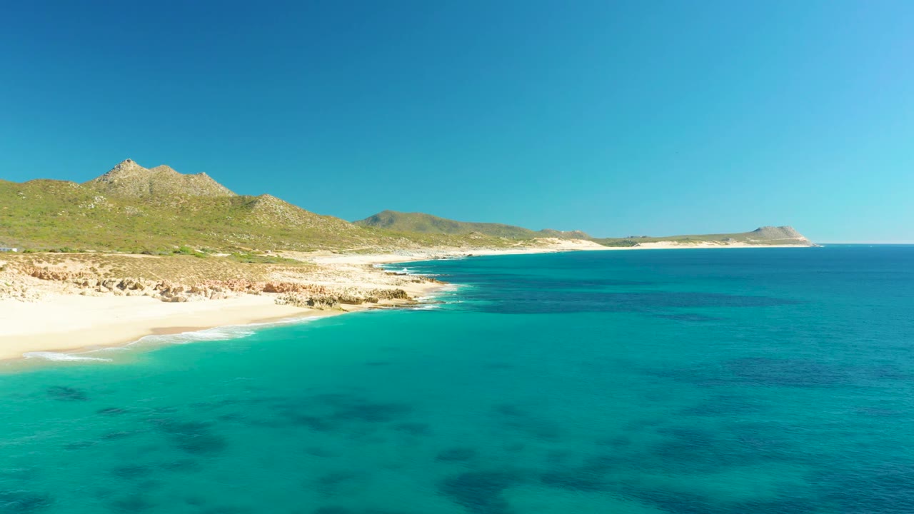 Opening frame of Buzzards Beach on the East Cape of Baja California Sur, with turquoise shallows, pale sand, rocky coast, green hills, distant headland, clear blue sky, and no visible people.