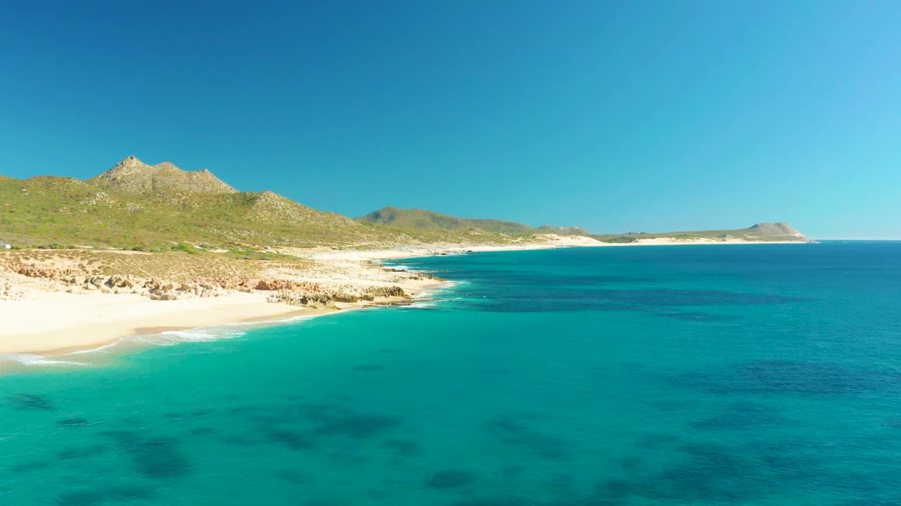 Buzzards Beach shoreline one second into the clip, showing blue-green water curling toward the sand, rocky outcrops, green hills, a distant point of land, and no visible people.
