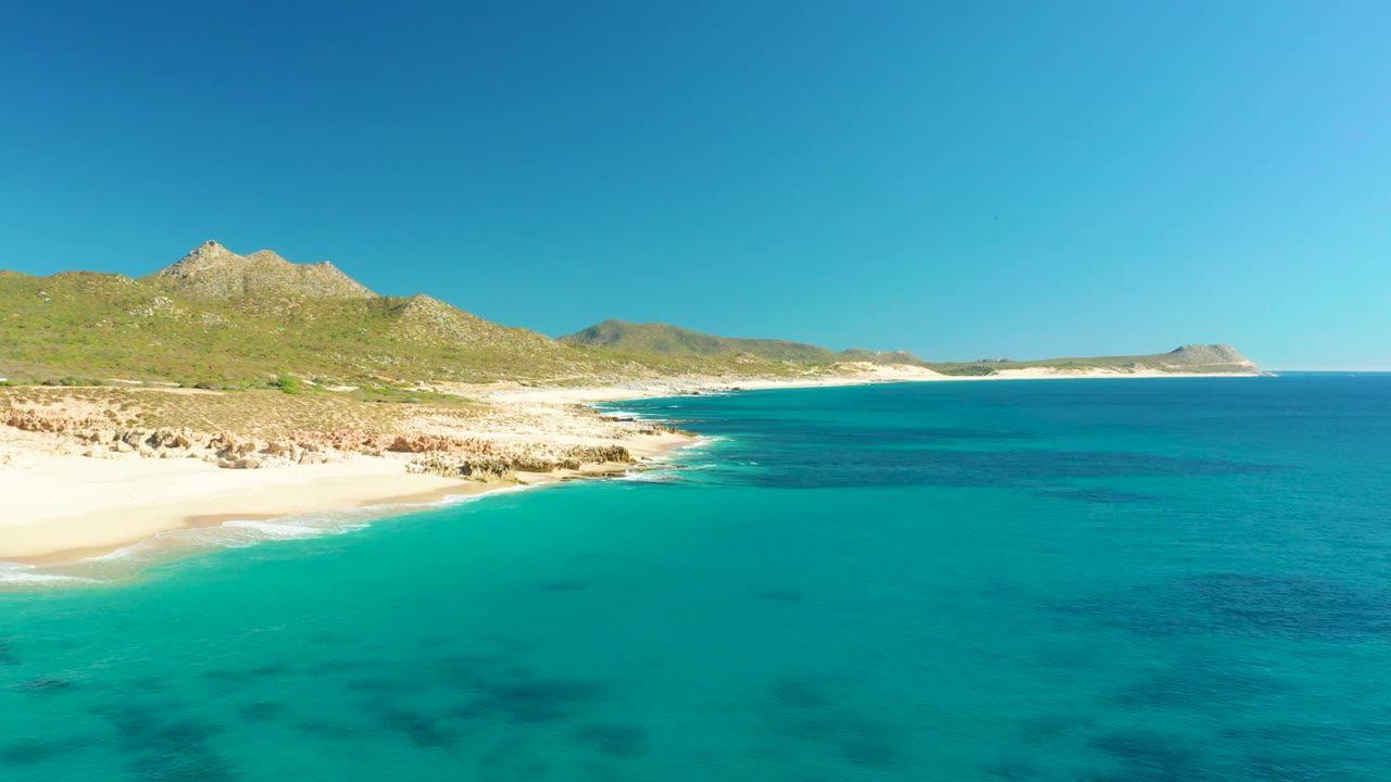 A wide view of Buzzards Beach in Baja California Sur, with turquoise shallows in the foreground, a sandy curve at left, rocky shoreline, low green hills, open sea, and no visible people.