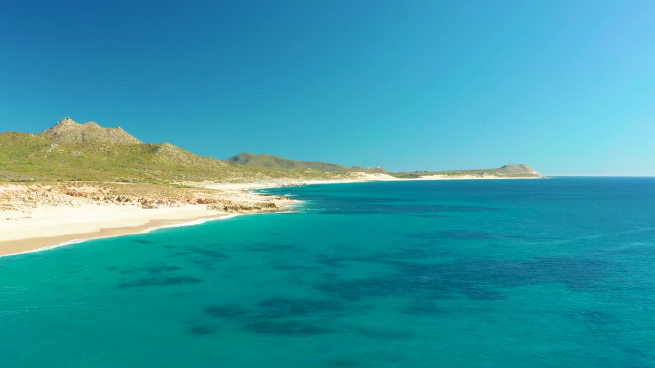Opening aerial frame over Buzzards Beach in Baja California Sur, with turquoise water in the foreground, pale sand at left, low mountains beyond the shore, and no visible people.