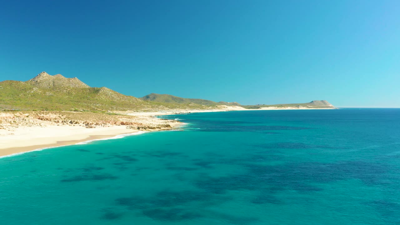 Turquoise water, sandy shoreline, rocky coast, and mountains under blue sky.