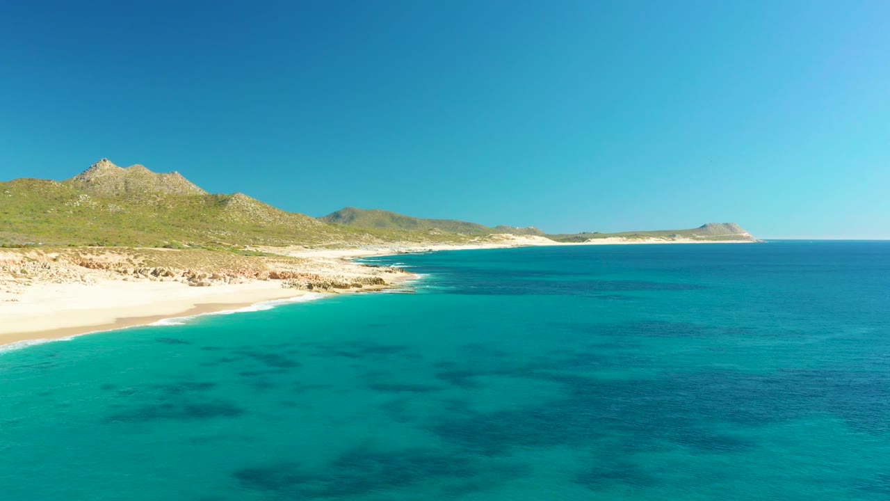 Aerial view of Buzzards Beach at 0:02, with blue-green ocean occupying most of the frame, pale beach and rocky points on the left, a low mountain ridge behind, and no visible people.