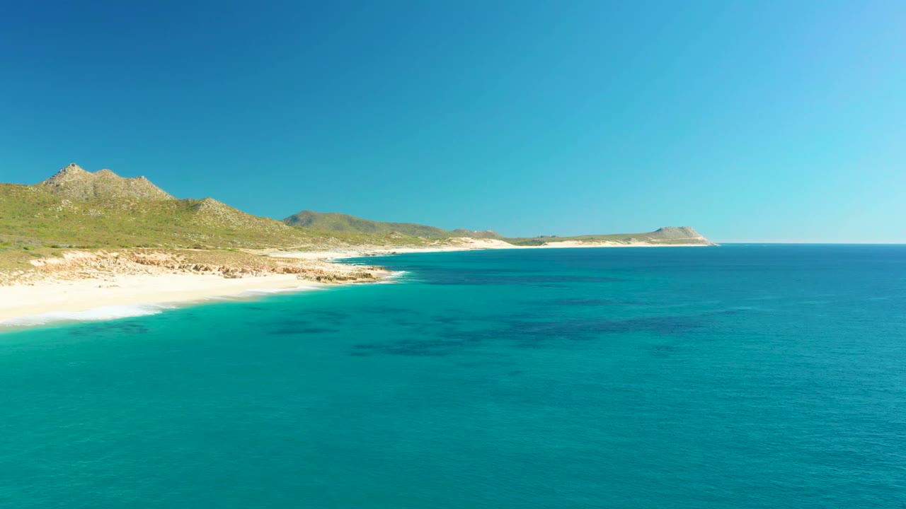 Opening aerial view of Buzzards Beach in Baja California Sur, with clear turquoise water in the foreground, pale sand along the left shore, green hills beyond, and no visible people.