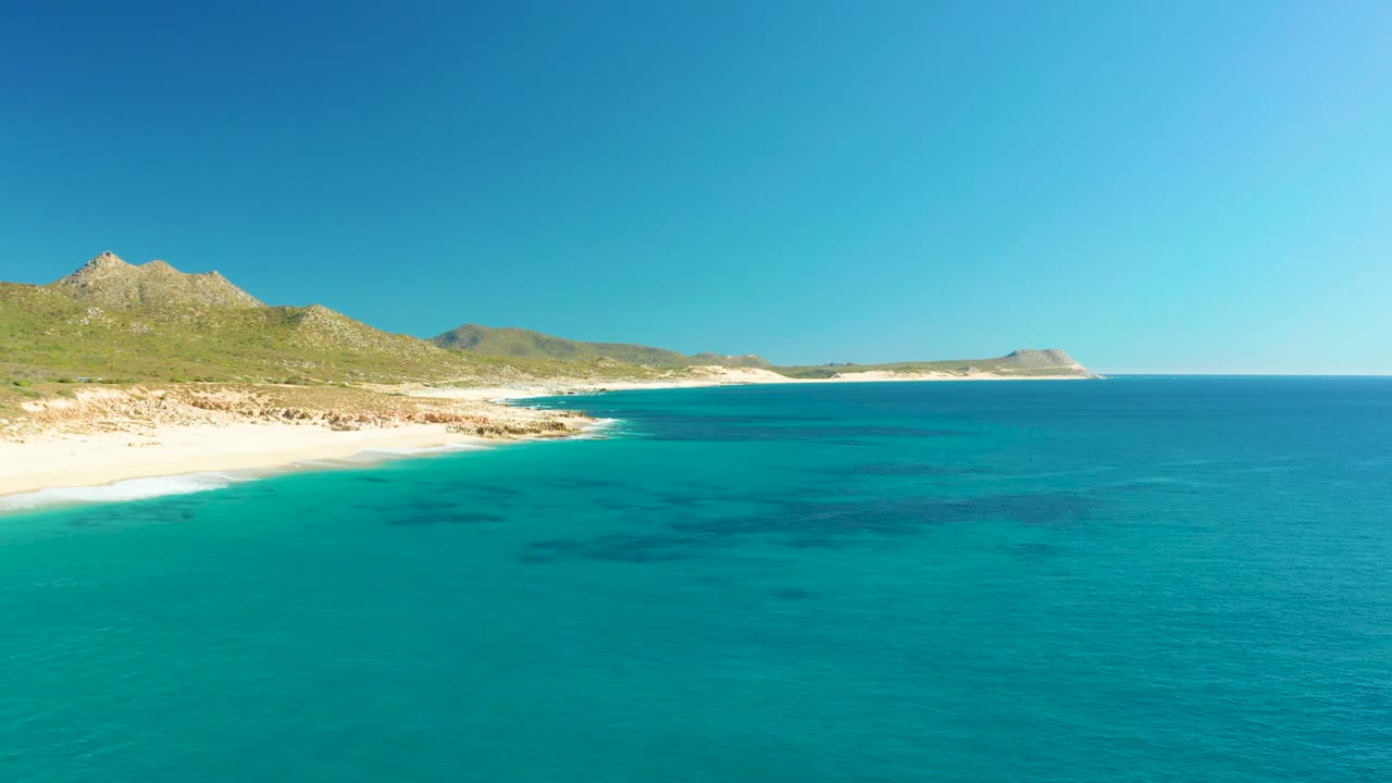 Clear blue water and curving sand beach below green hills.