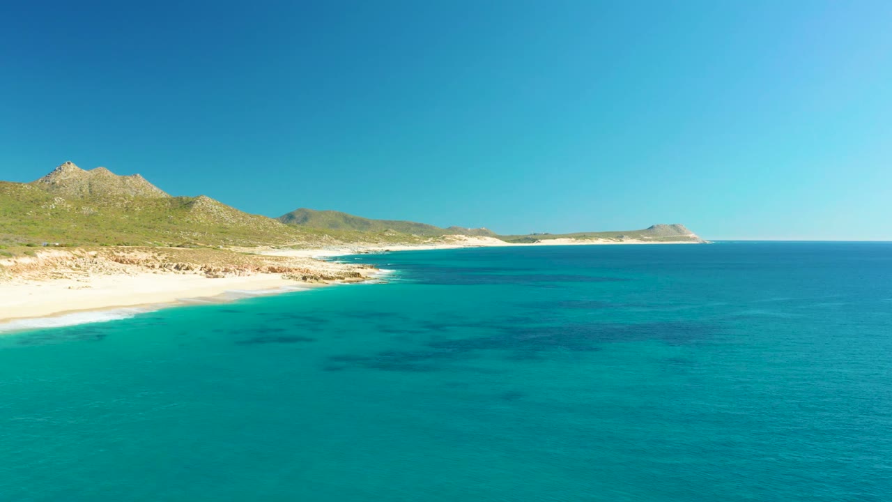 Wide aerial view over Buzzards Beach with turquoise shallows, darker underwater patches, pale sand at left, low hills behind the shore, and no visible people.