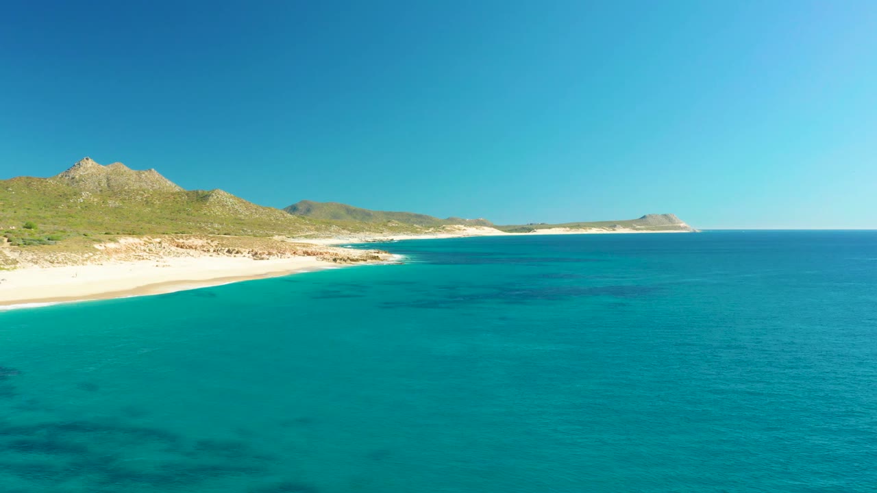 Opening frame of Buzzards Beach East Cape in Baja California Sur, with turquoise water in the foreground, sandy beach at left, green hills behind the shore, and no visible people.
