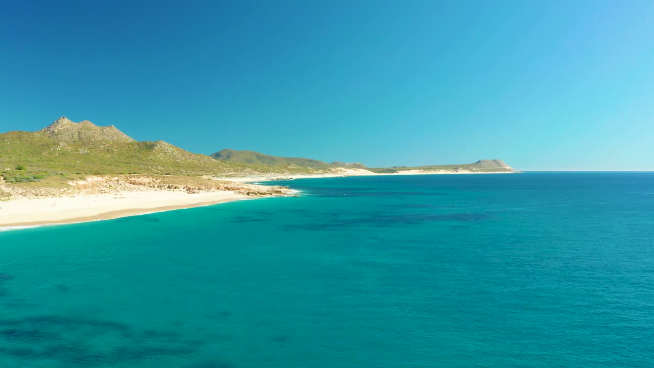 Wide coastal view with turquoise sea, pale beach, green hills, and distant headland.