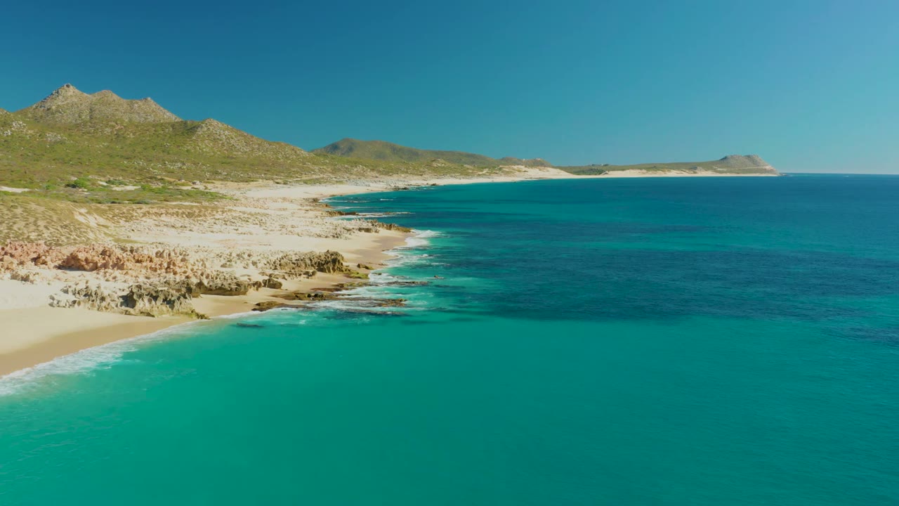 Opening frame over Buzzards Beach East Cape, Mexico, with turquoise water filling the foreground, pale sand and rocks along the left shoreline, low hills in back, and no visible people.