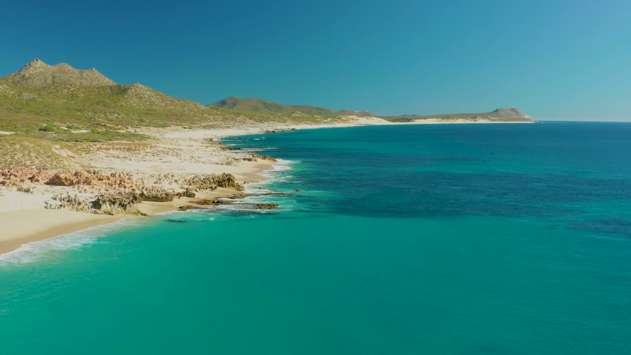 Aerial coastline with beach rocks, turquoise ocean, hills, and distant headland.