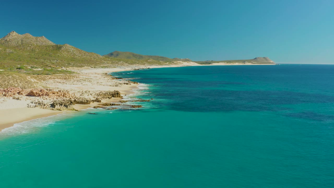 At 0:02, the aerial view shows more open turquoise water in front of the Buzzards Beach sand and rocky edge, with hills stretching across the background and no visible people.