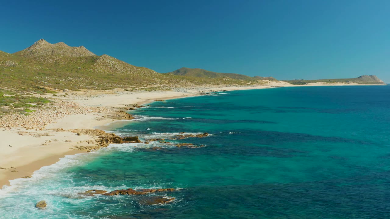 Buzzards Beach East Cape shoreline in Baja California Sur with turquoise water, tan sand, rocky surf, low mountains, clear blue sky, and no visible people at the opening moment.