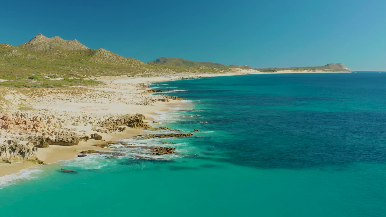 Aerial view of Buzzards Beach East Cape in Baja California Sur, with turquoise water in the foreground, sandy rocky shore at left, low hills beyond, and no visible people.