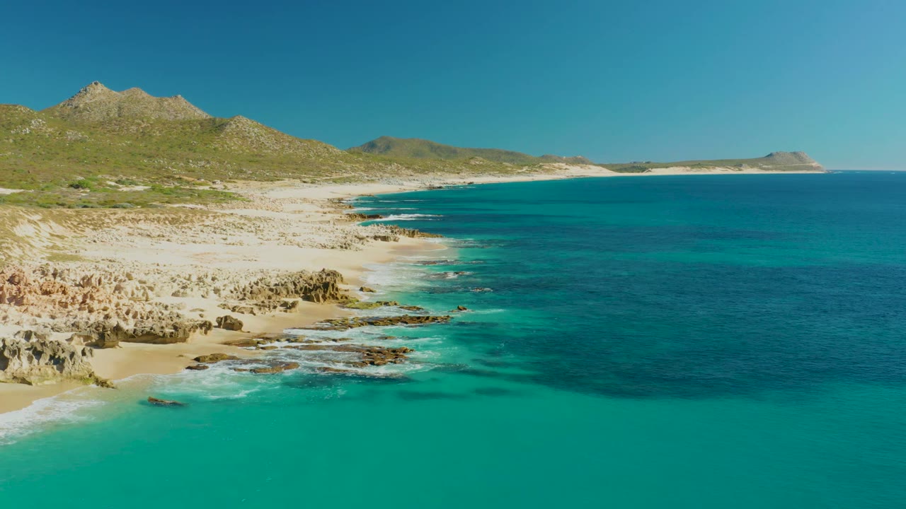 Wide aerial coastline with teal water, pale sand, rocks, and hills under clear sky.